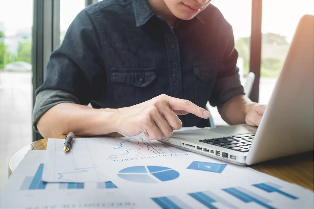 Analyst comparing printed compliance reporting against figures on a laptop